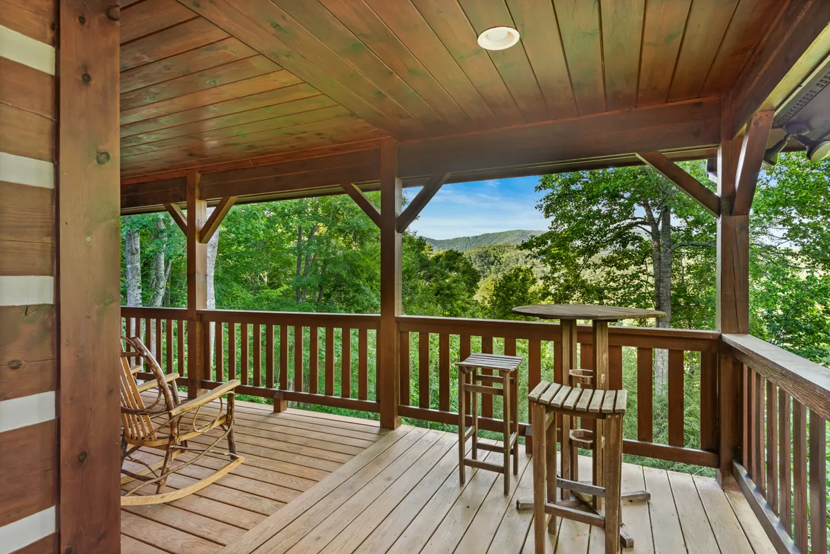 The image shows a serene covered deck with wooden railing, classic rocking chair, and bar stools. Beyond the deck, sweeping mountain views stretch into the distance, creating an inviting space for quiet reflection, conversation, or enjoying a book while soaking in the scenery.