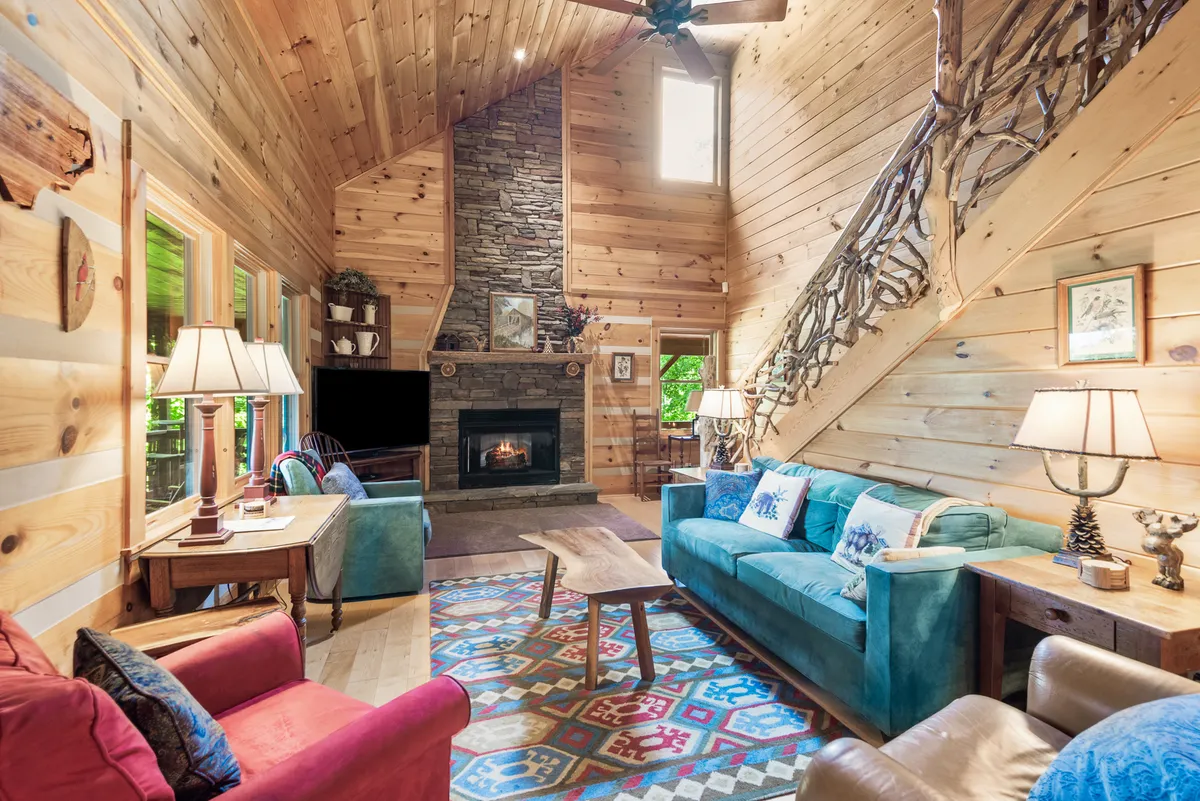 A dramatic living area with a striking floor-to-ceiling stone fireplace, a colorful area rug, wooden ceiling and walls, and large windows that frame panoramic mountain views. Plush seating invites relaxation and connection.