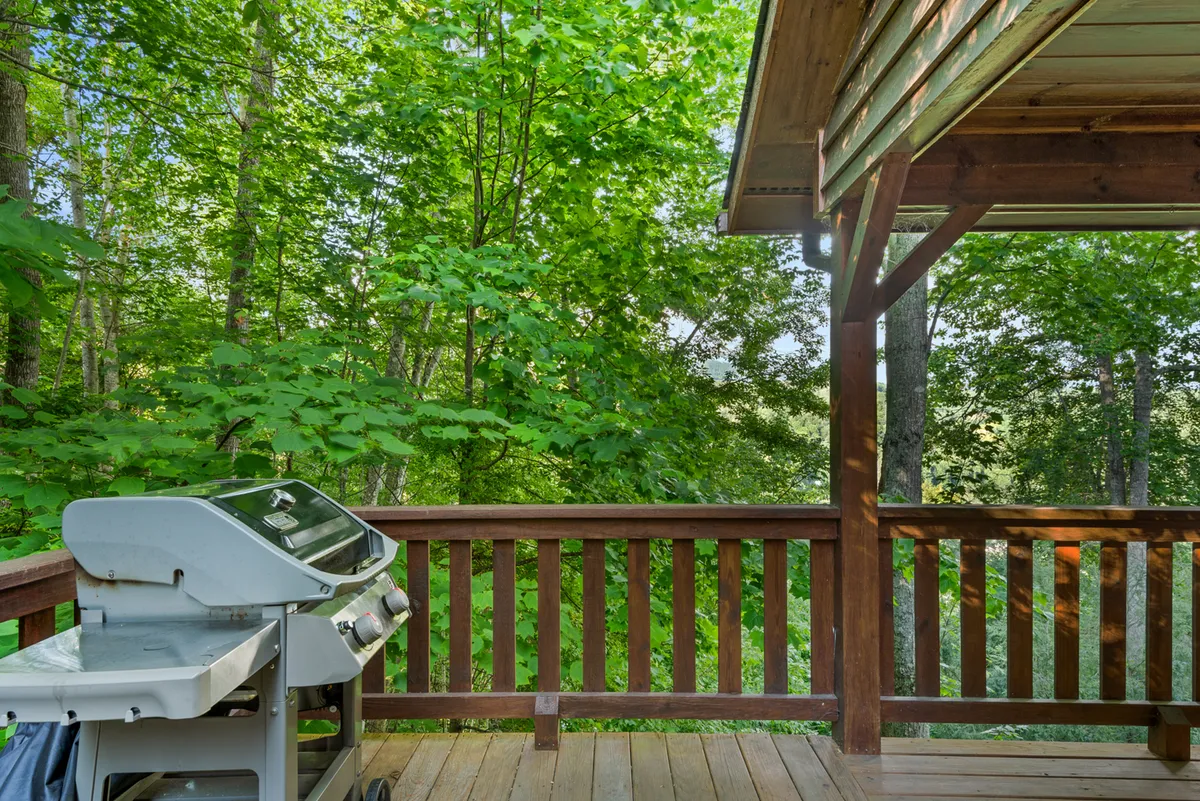 An exterior deck scene featuring a grill, wooden railing, and surrounding greenery. The space is set up for outdoor dining and socializing, ideal for gathering with friends and family to share stories and meals outdoors.