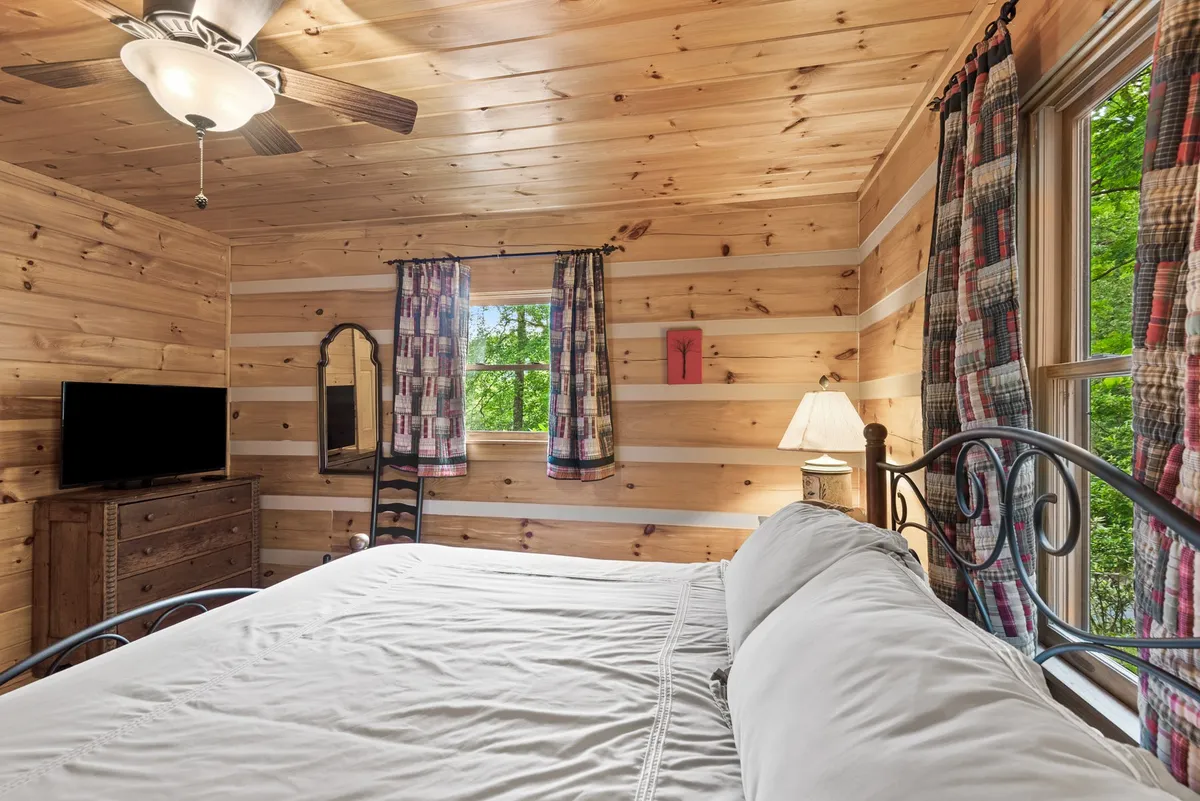 This image features a serene bedroom with a bed dressed in white linens, a wooden dresser, a large window with a view, and soft curtains. The mood is one of inviting tranquility, emphasizing morning sunlight and relaxation while experiencing the natural surroundings from indoors.