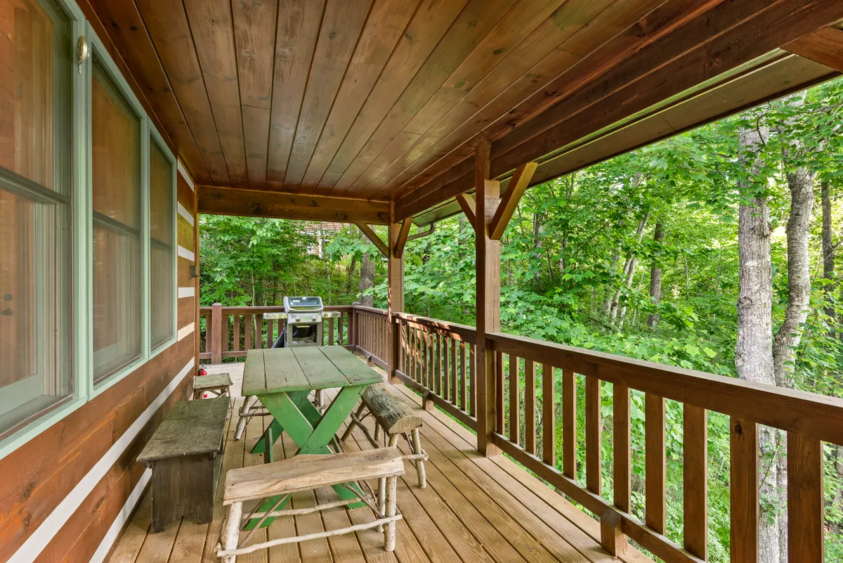 This view features a spacious wooden deck equipped with a green table for al fresco dining, a barbecue grill, and is shaded by surrounding trees. The atmosphere is cozy and relaxed, ideal for family gatherings or meals outdoors.