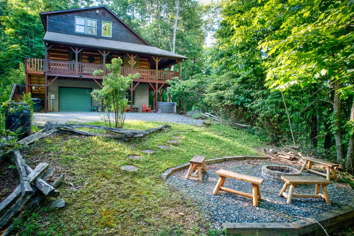 The scene centers on a fire pit area with wooden benches and a spacious patio surrounded by trees, with the house in the background. The fire pit area is set up for cozy gatherings under the stars, perfect for evening s’mores, stories, and celebrations.