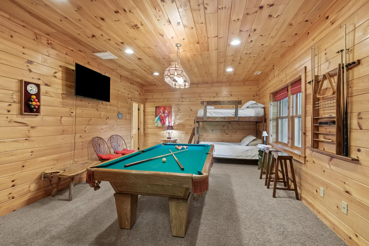 This interior image features a recreation room with a pool table in the foreground, cozy bunk beds, wooden walls, and a chandelier overhead. The space is clearly designed for play and relaxation, with plentiful space for group activities and lounging.