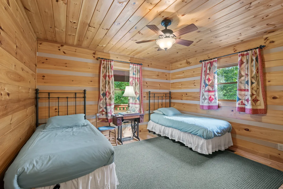 A cozy bedroom featuring two twin beds with light blue bedding, wood-paneled walls and ceiling, colorful curtains with patchwork designs, and a small nightstand with a lamp. The window streams in daylight, giving the room a cheerful, inviting vibe—perfect for children or extra guests.
