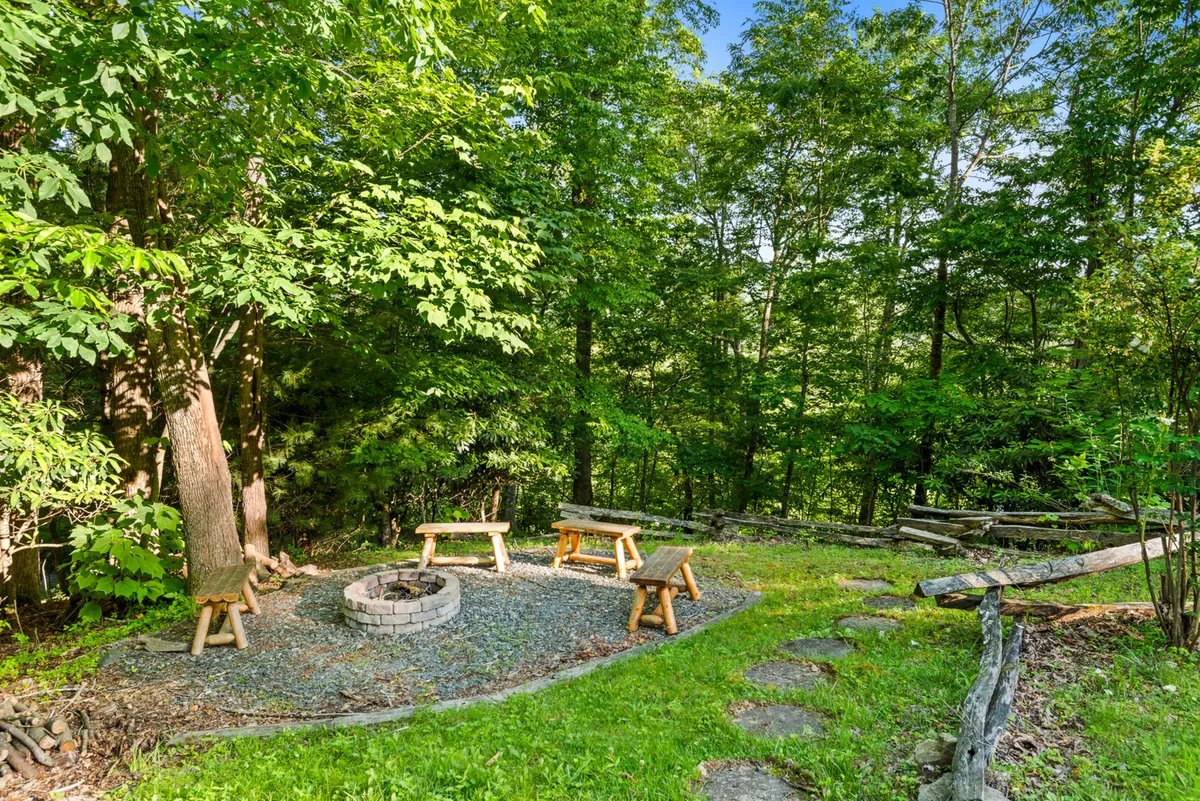 This image shows a cozy outdoor fire pit surrounded by wooden benches, set among tall trees. The natural setting makes it ideal for gathering in the evening, roasting marshmallows, telling stories, and looking up at the night sky.