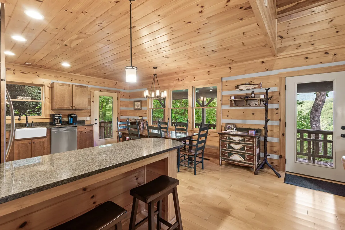 Highlighting a dining area with a granite countertop, rustic wooden dining table, and large windows that frame the outdoors, this image showcases a space for shared meals and quiet moments. The abundance of natural light and the proximity to nature are evident, reinforcing the sense of being nestled in the mountains.
