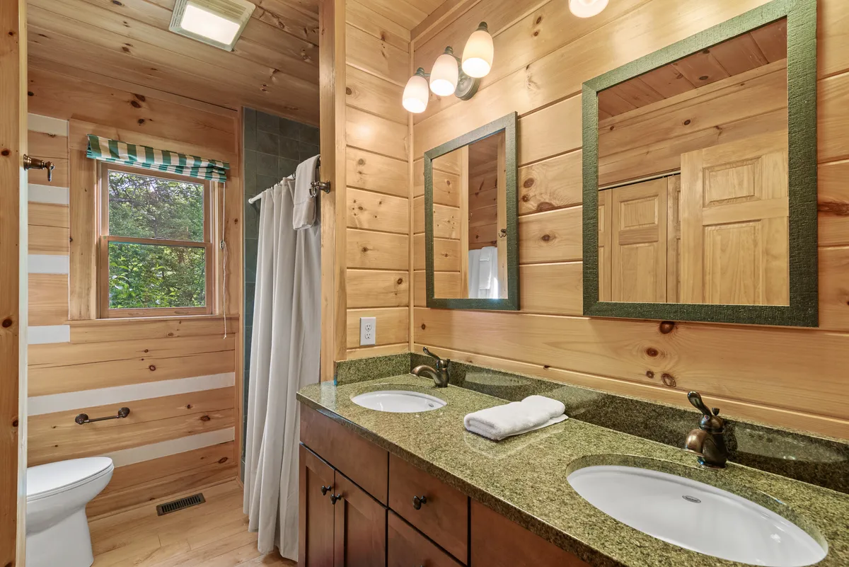 A spa-inspired bathroom with double sink vanity, large mirrors, wood-paneled walls, a shower curtain, and a window with a peaceful view. The space exudes warmth, comfort, and functionality, with plush towels and natural light enhancing the relaxing atmosphere.